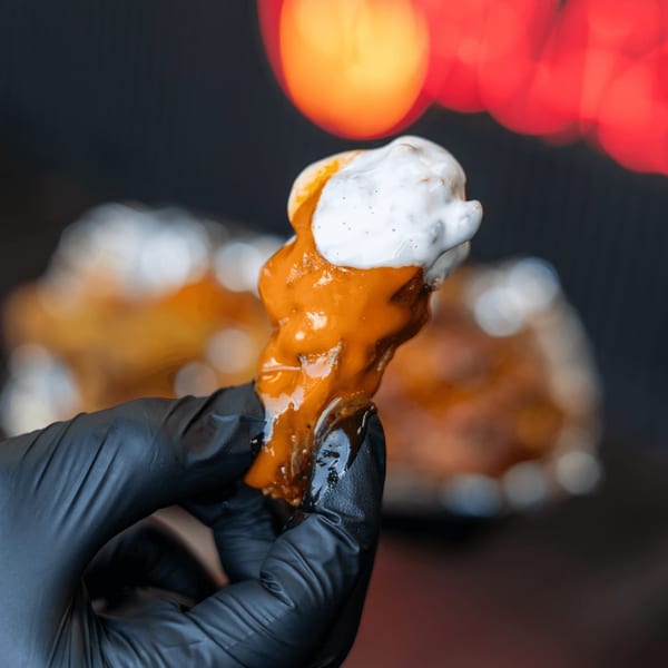 A gloved hand holds a Blazin’ Grill chicken wing, dripping with orange sauce and white dressing.