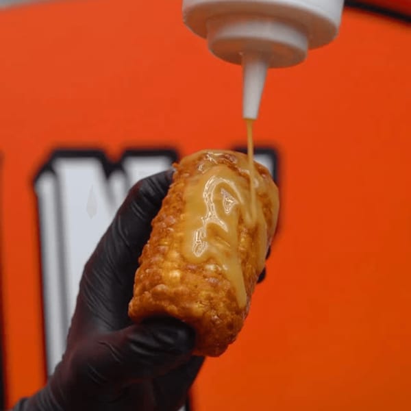A gloved hand holds a crispy Blazin’ Grill corn dog as cheese sauce is drizzled over it.