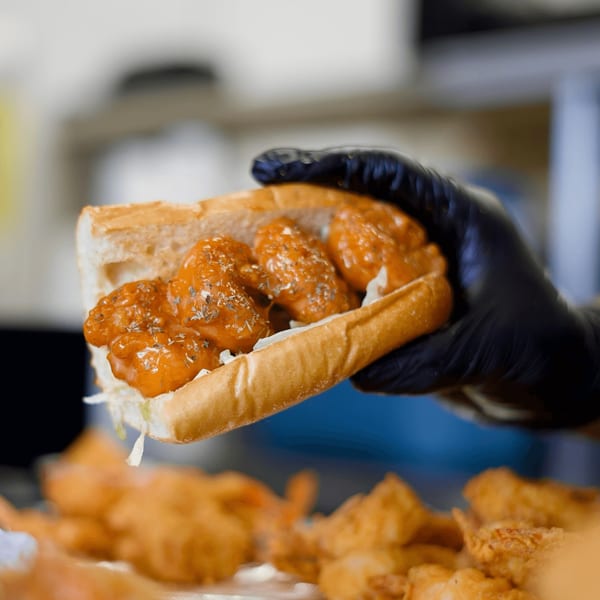 A gloved hand holds a Blazin' Grill shrimp po'boy sandwich with orange sauce and herbs.